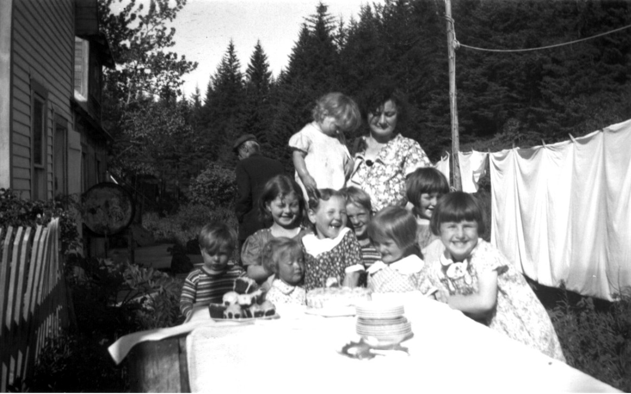 Table with a cake and candles, 8 children crowded together at the end, behind them a woman in a print dress holding a child looking down; house to the left, laundry hung on a line to the right
