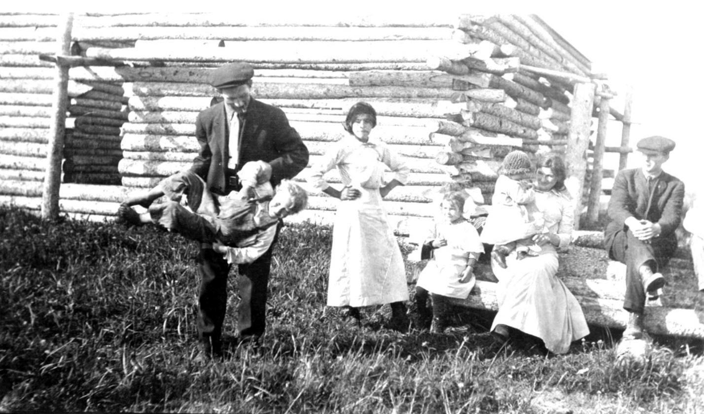 Grandparents sitting beside a log cabin with the author's mother, while a woman and a girl watch a man swinging a boy