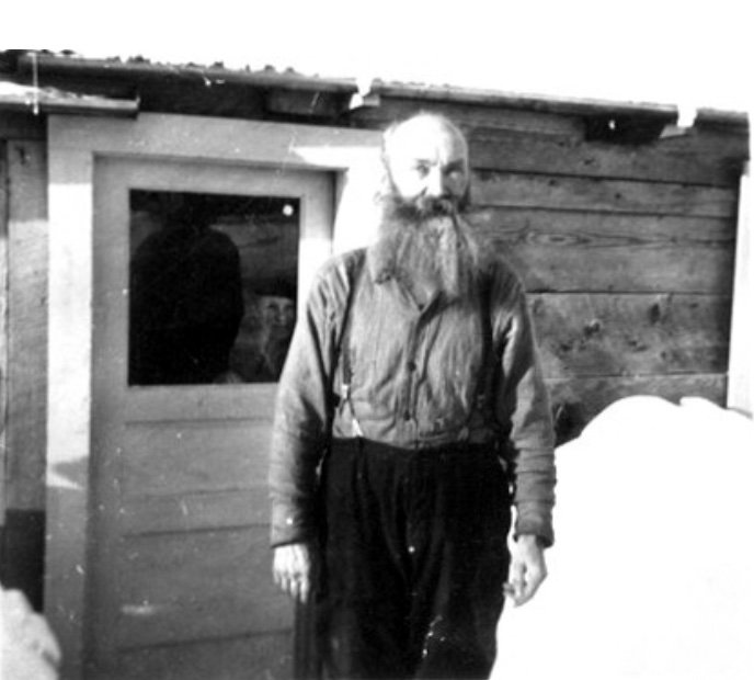 Bald man with full beard, suspenders, standing before the door of an unpainted house and a pile of snow