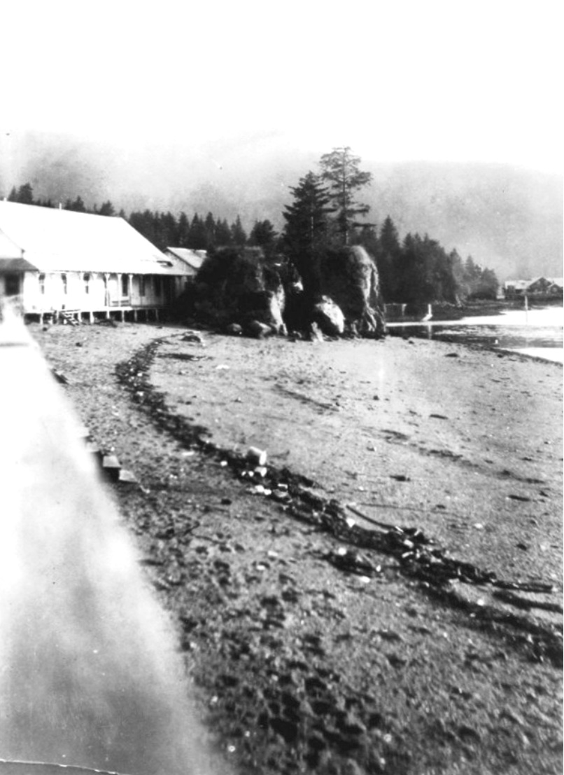 Building on posts at the left on a graveled beach, large rocks at the center, and in the distance the old cannery and hill