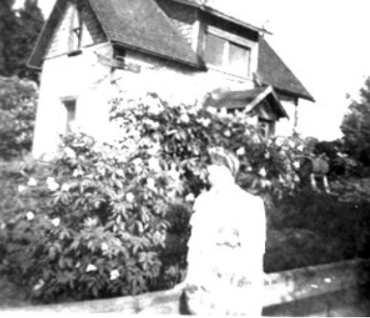 Woman standing before two-storey house, flowering bushes obscuring the first floor