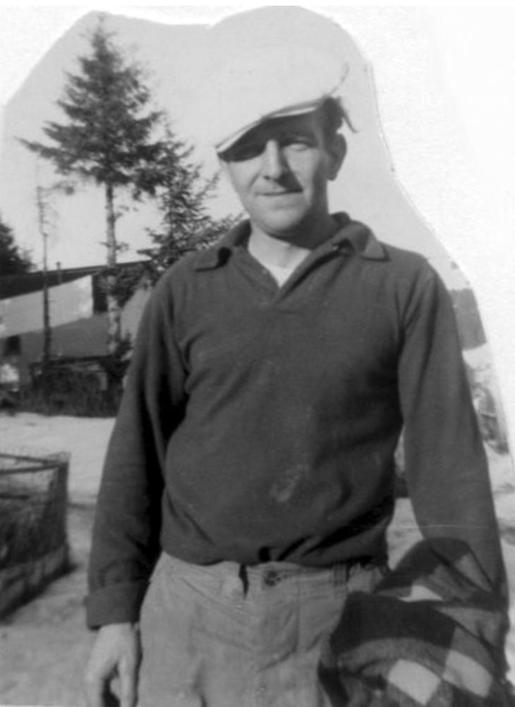 Man holding a wool jacket wearing a driving cap. Behind is a pruned pine tree by a building.