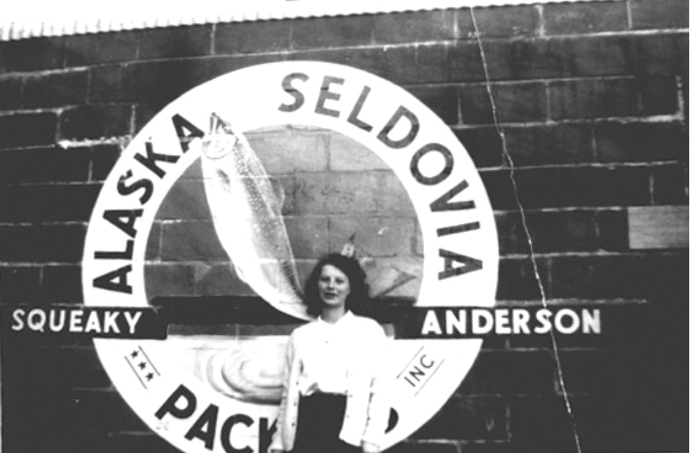 The author (age 17) standing before a cinderblock wall painted with a large circle featuring a leaping salmon, around the circle ALASKA SELDOVIA PACKERS INC, across the circle SQUEAKY ANDERSON