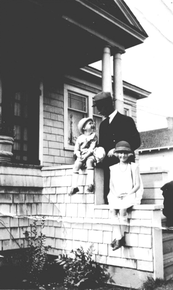 Man in suit and hat looking down on a small boy sitting with his sister on the rails of front steps