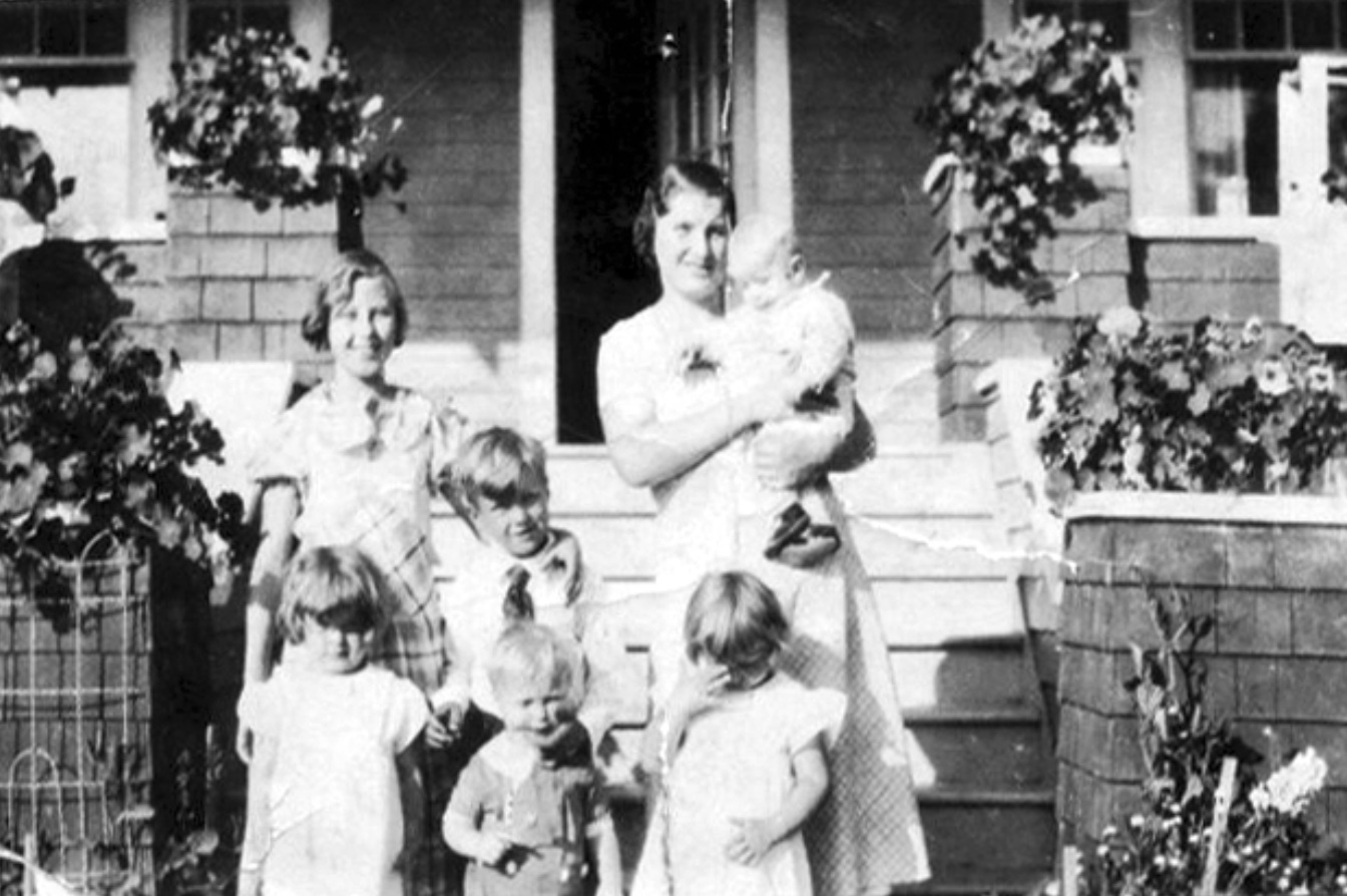 Woman carrying a baby and posing with four young children and an older girl at the foot of steps to a house