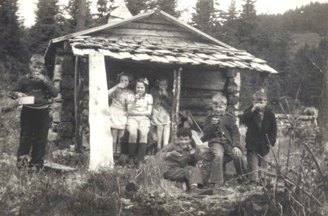 Seven children at the end of a small log cabin, all drinking something