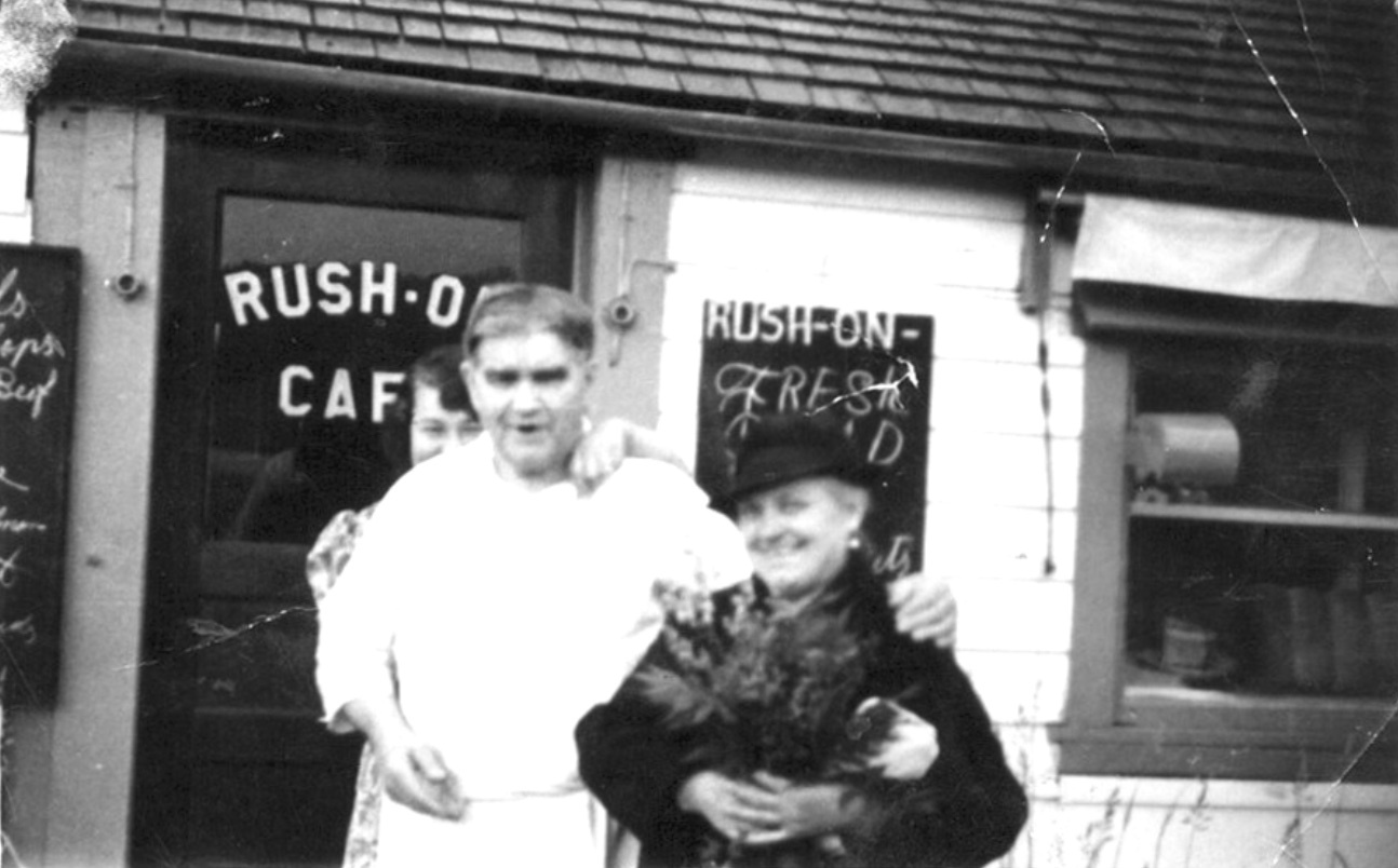 Man in a white chef’s suit, a woman peering over his right shoulder with her hand on his left shoulder, his hand on a shorter woman’s shoulder, dressed in black with a black hat and a floral arrangement in her arms; behind them, a door painted RUSH-ON CAFE, and a chalk board advertizing FRESH BREAD