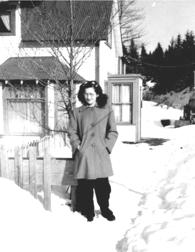 Young woman standing in snow wearing pants, overcoat, and glasses outside a house