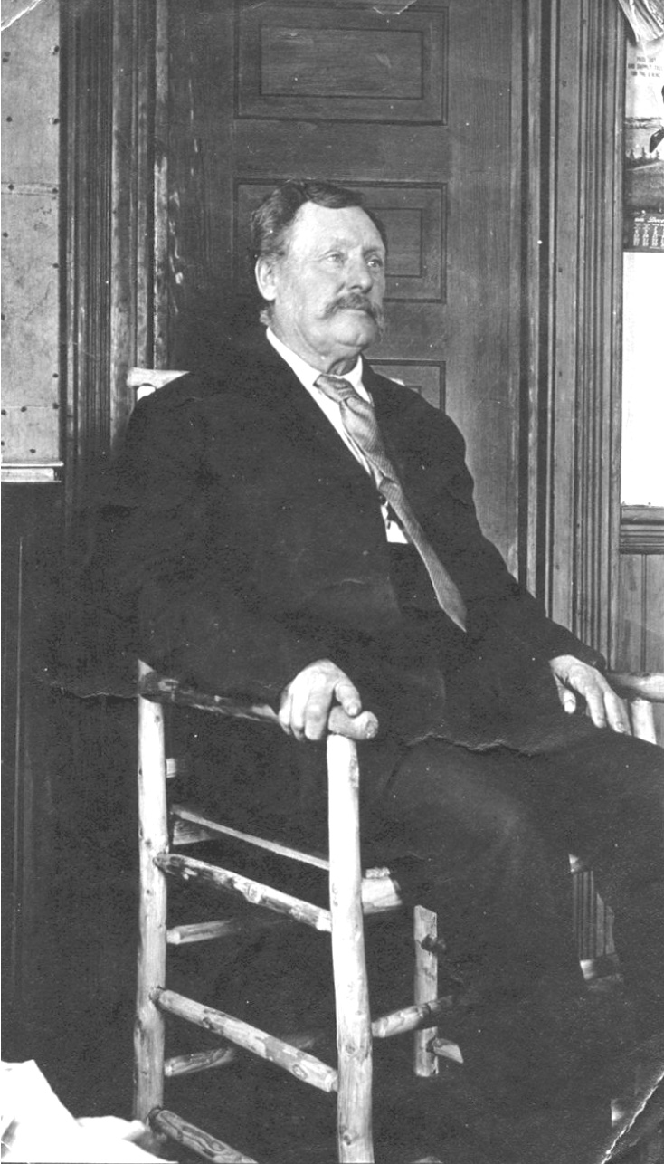 Man with handlebar mustache, in suit and tie, seated on a rough handmade chair before a door
