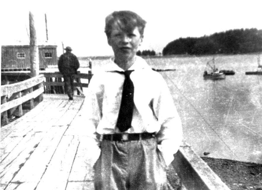 A boy on the boardwalk wearing a tie, hands in his pockets. The bay with fishing boats are to his left