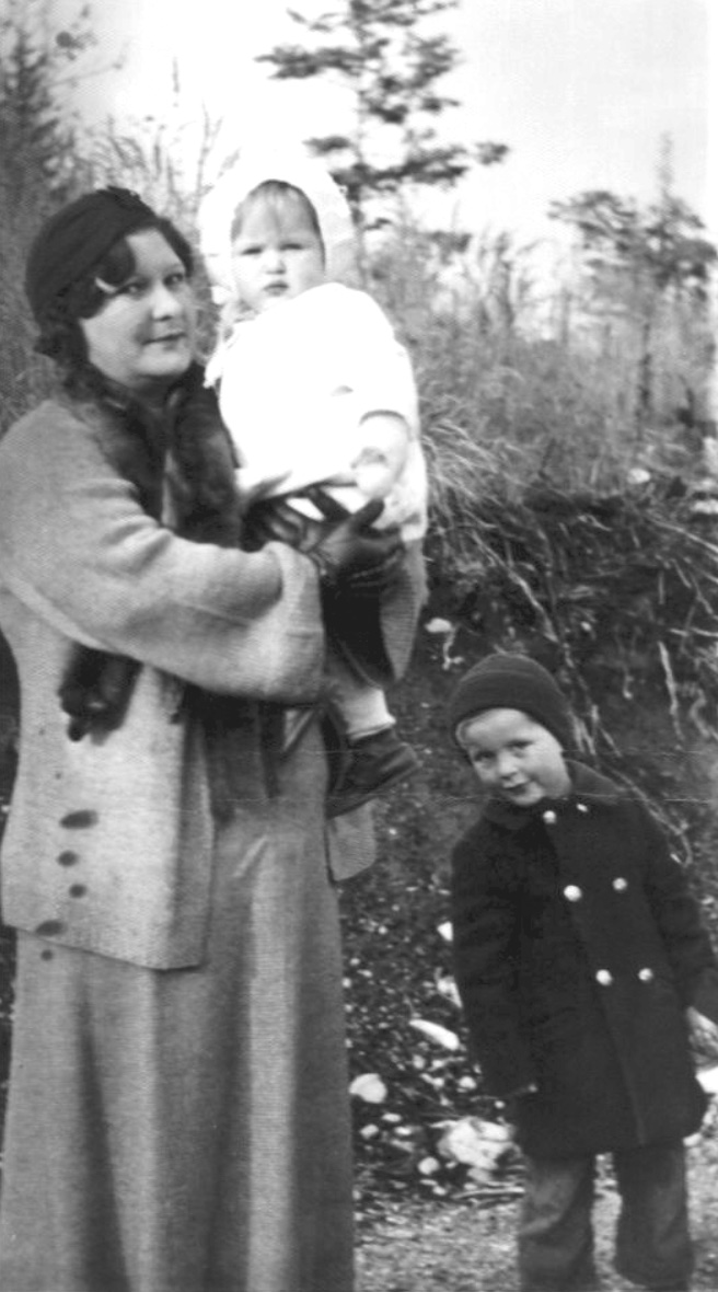 Woman wearing sweater and scarf holding a baby, a boy in overcoat and knit hat leans over and smiles