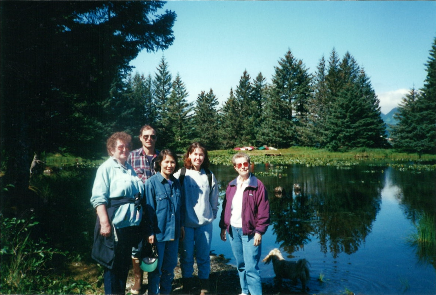 Four women and a man standing together before a small lake; fir trees on the other side of the lake