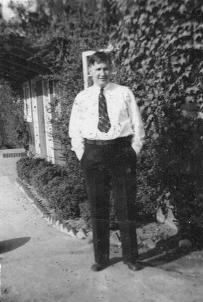 Man in dress shirt and tie, hands in pockets, standing beside ivy-covered wall outside a house.