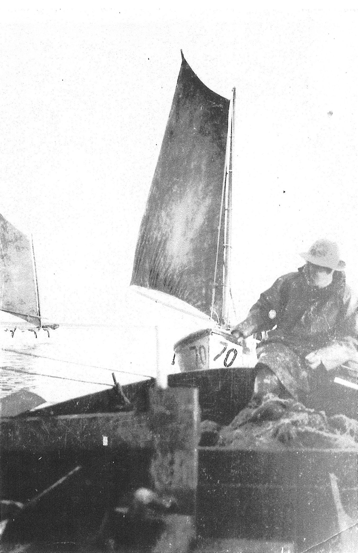 Man on a small boat wearing a rain slicker and hat; a small sailboat behind him is labeled 70.
