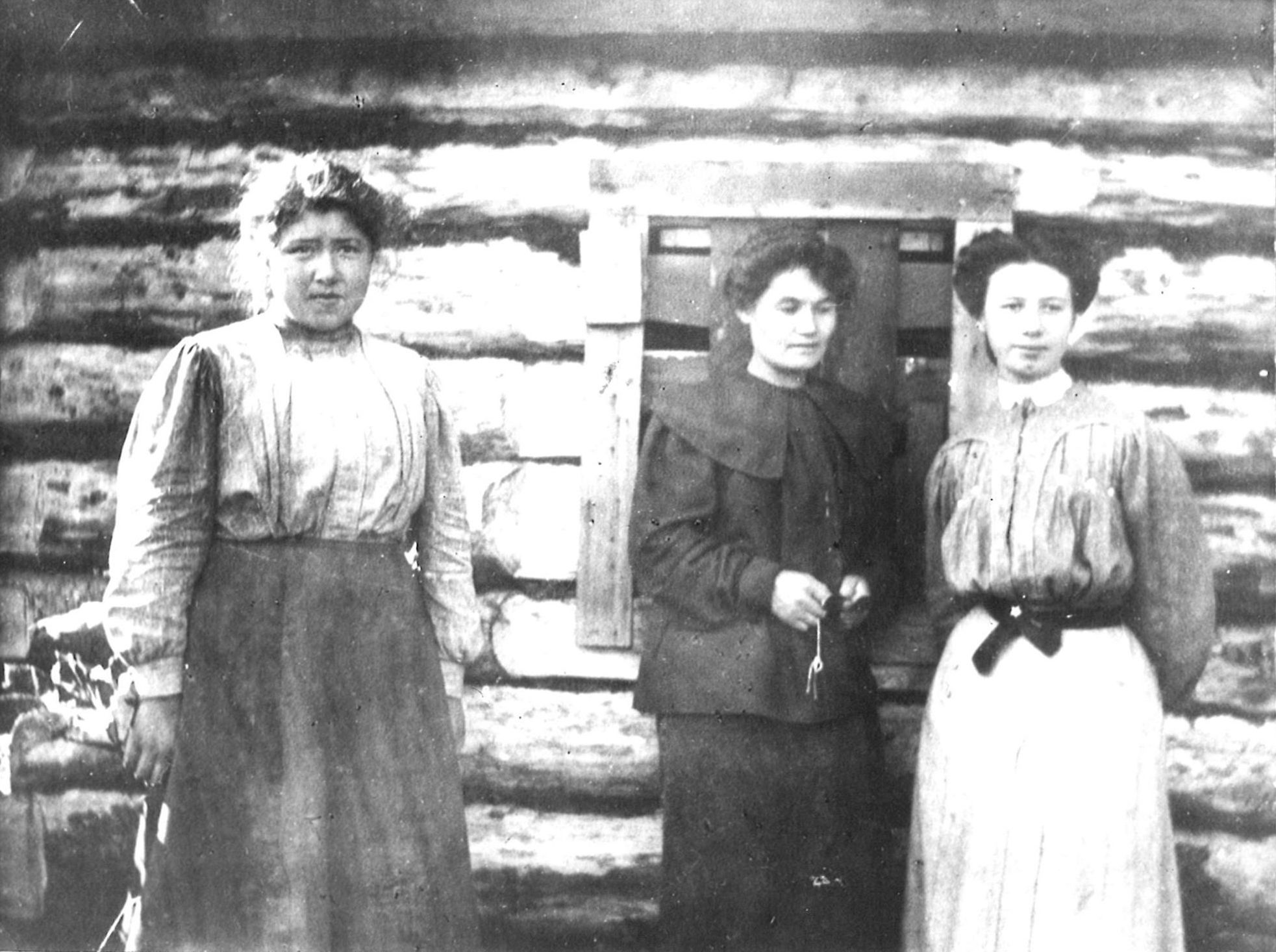 Three women standing before a log house