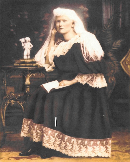 A young woman with long white hair in a dress with lace collar sits at a table with a book in her lap