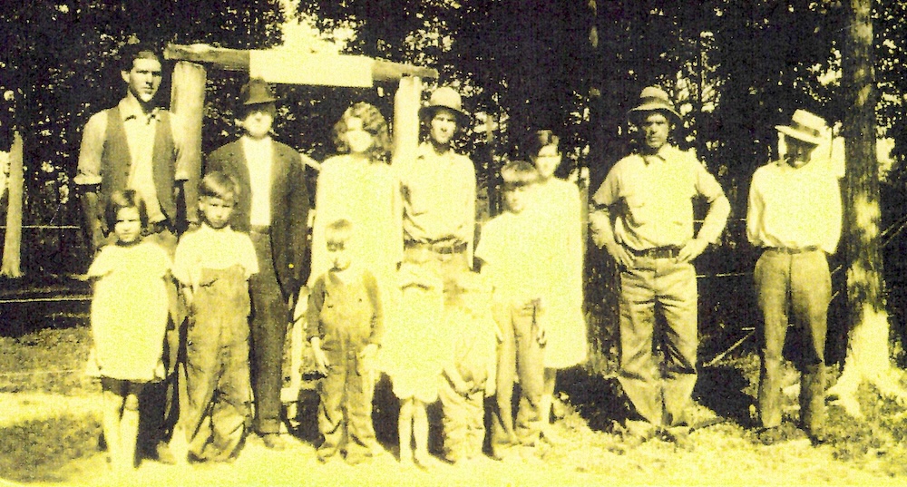 Group photo of adults and children before a gate for a bobwire fence, trees behind them