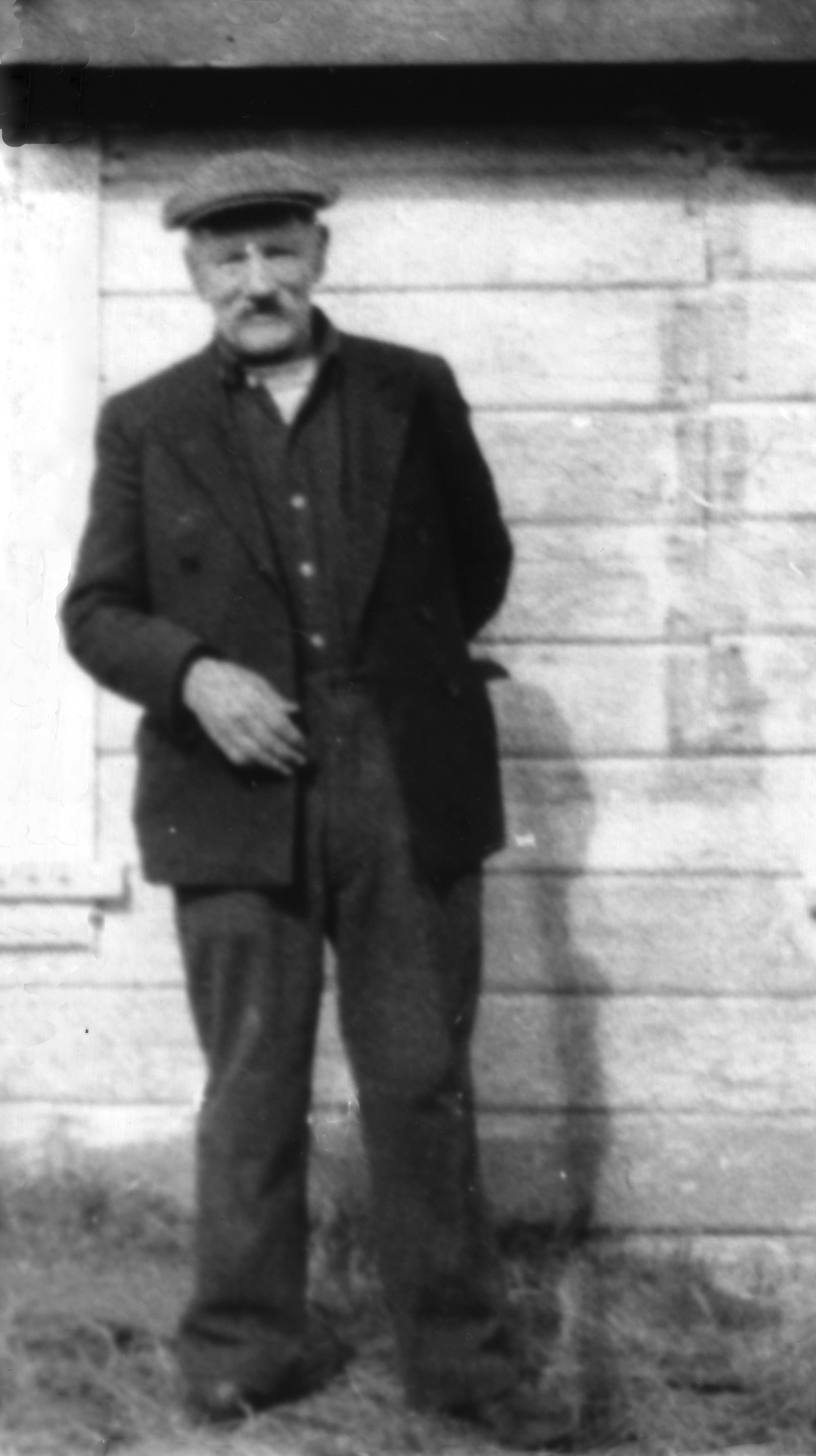 Man with mustache standing before a house in overcoat and cap, right hand in pocket